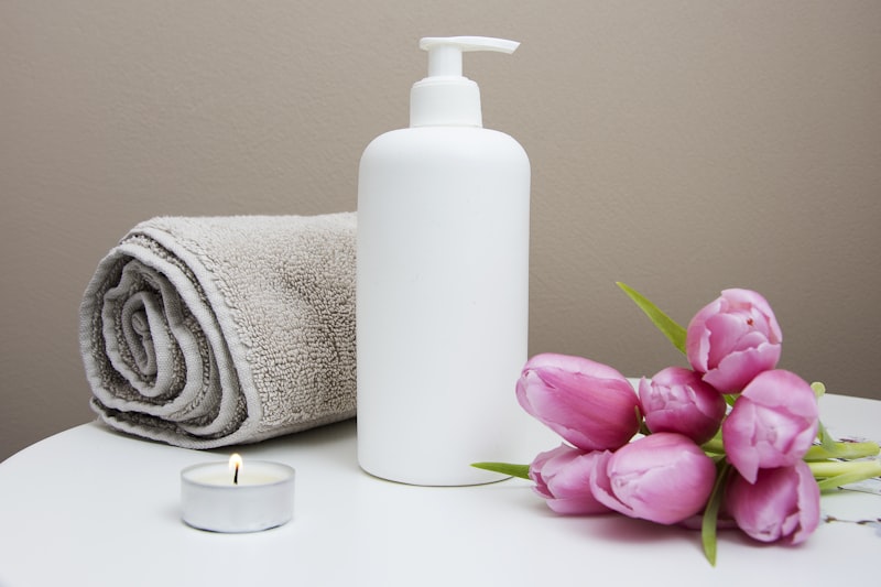 At-home spa setup with white pump bottle, rolled towel, lit candle, and pink tulips